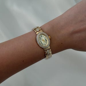 May include: A gold-tone oval wristwatch featuring a bezel encrusted with clear crystals. The watch face has a cream background with black Roman numeral hour markers and gold hands. The metal link bracelet is also accented with small clear crystals.