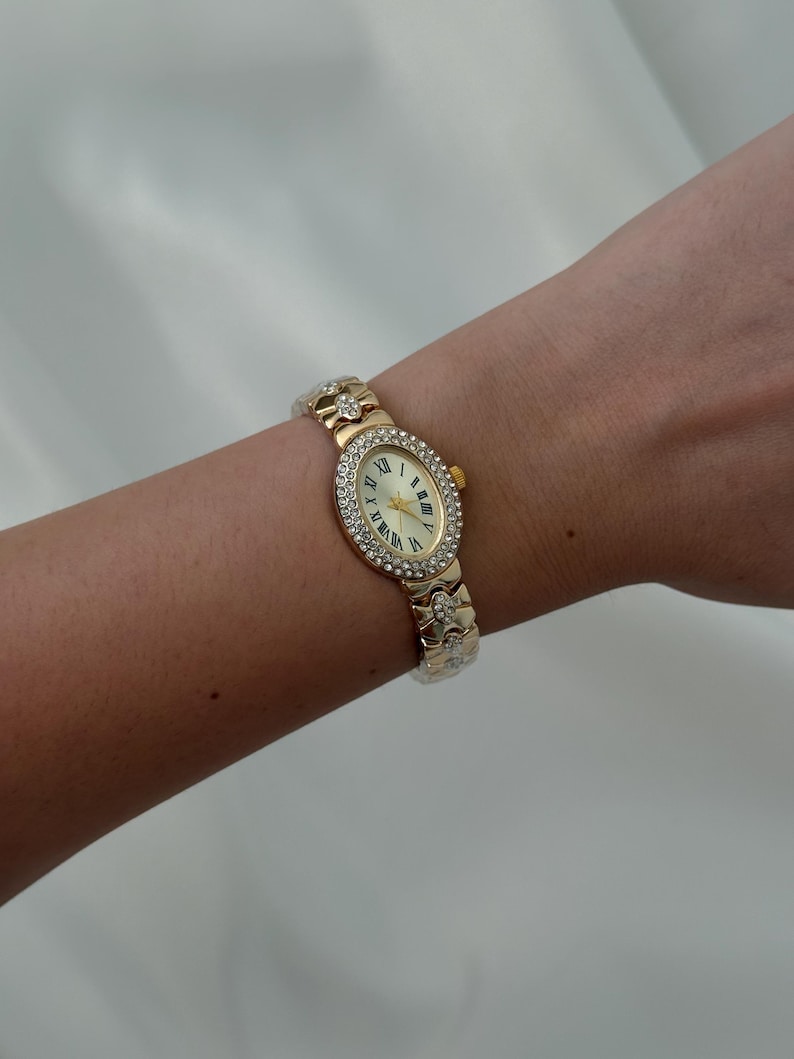 May include: A gold-tone oval wristwatch featuring a bezel encrusted with clear crystals. The watch face has a cream background with black Roman numeral hour markers and gold hands. The metal link bracelet is also accented with small clear crystals.