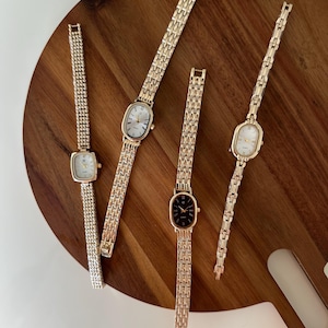 Puede incluir: Vista cenital de cinco relojes de pulsera dorados con diferentes formas de esfera: cuadrada, ovalada y rectangular. Las esferas son blancas o negras, con agujas y marcadores dorados. Los relojes están sobre una superficie de madera.