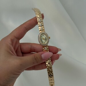May include: A gold-tone wristwatch featuring an oval-shaped face with a white dial and Roman numerals. The bezel and select links on the bracelet are embellished with clear crystals. The watch has a link-style band and a small crown on the side.