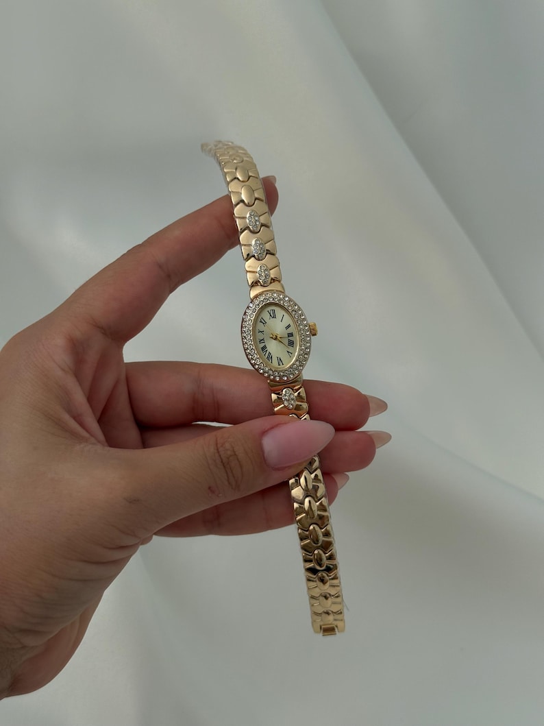 May include: A gold-tone wristwatch featuring an oval-shaped face with a white dial and Roman numerals. The bezel and select links on the bracelet are embellished with clear crystals. The watch has a link-style band and a small crown on the side.