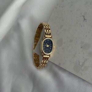 Puede incluir: Reloj de pulsera dorado con esfera ovalada negra y agujas doradas. El reloj tiene una correa de eslabones y un movimiento de cuarzo. El reloj se muestra sobre una superficie de sat&eacute;n blanco.