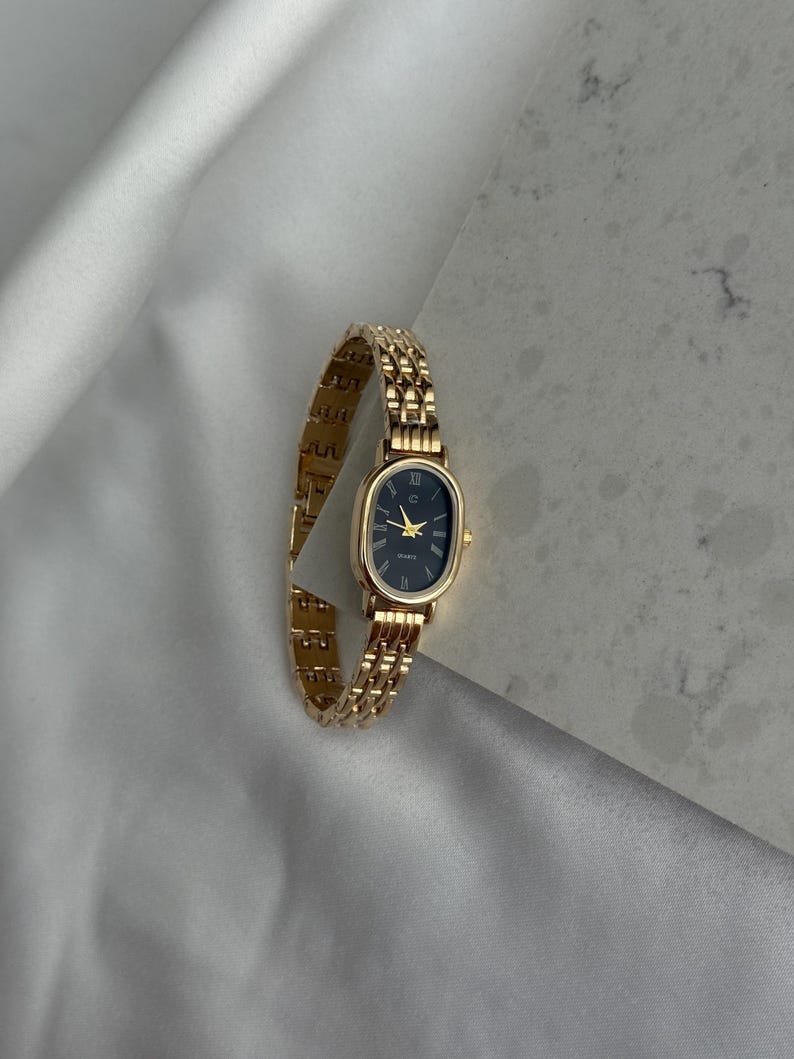 Puede incluir: Reloj de pulsera dorado con esfera ovalada negra y agujas doradas. El reloj tiene una correa de eslabones y un movimiento de cuarzo. El reloj se muestra sobre una superficie de sat&eacute;n blanco.