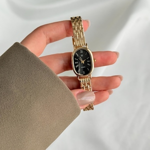 Puede incluir: Un reloj de cuarzo dorado con esfera ovalada negra y agujas doradas. El reloj tiene una correa de eslabones dorados. El reloj se sostiene sobre un fondo blanco.