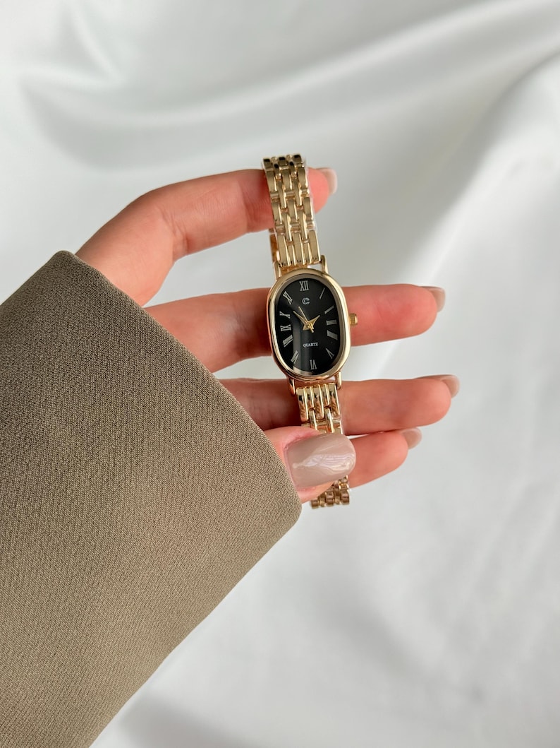 Puede incluir: Un reloj de cuarzo dorado con esfera ovalada negra y agujas doradas. El reloj tiene una correa de eslabones dorados. El reloj se sostiene sobre un fondo blanco.