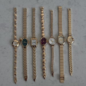 May include: Seven gold-tone wristwatches with various link styles and dial colors. The watches feature diverse face shapes including round, oval, and rectangular, with metallic bands ranging from delicate heart-shaped links to solid rectangular segments.