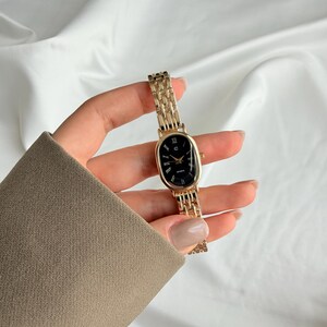 Puede incluir: Reloj de pulsera dorado con esfera ovalada negra y n&uacute;meros romanos. El reloj tiene una correa de eslabones y se sostiene sobre un fondo blanco. Un accesorio cl&aacute;sico.