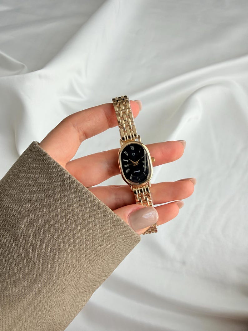 Puede incluir: Reloj de pulsera dorado con esfera ovalada negra y n&uacute;meros romanos. El reloj tiene una correa de eslabones y se sostiene sobre un fondo blanco. Un accesorio cl&aacute;sico.