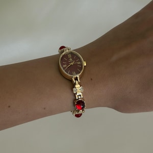 May include: A gold-tone wristwatch featuring an oval-shaped case with a dark red dial and Roman numeral markers. The bracelet is composed of gold-tone links interspersed with small clear crystals and round, deep red translucent cabochon stones.
