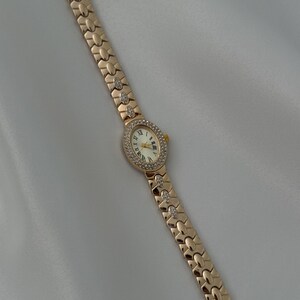 May include: A rose gold-tone wristwatch featuring an oval-shaped face with a white dial, black Roman numeral hour markers, and gold-tone hands. The bezel and select links on the metal bracelet are embellished with small, clear, sparkling stones.