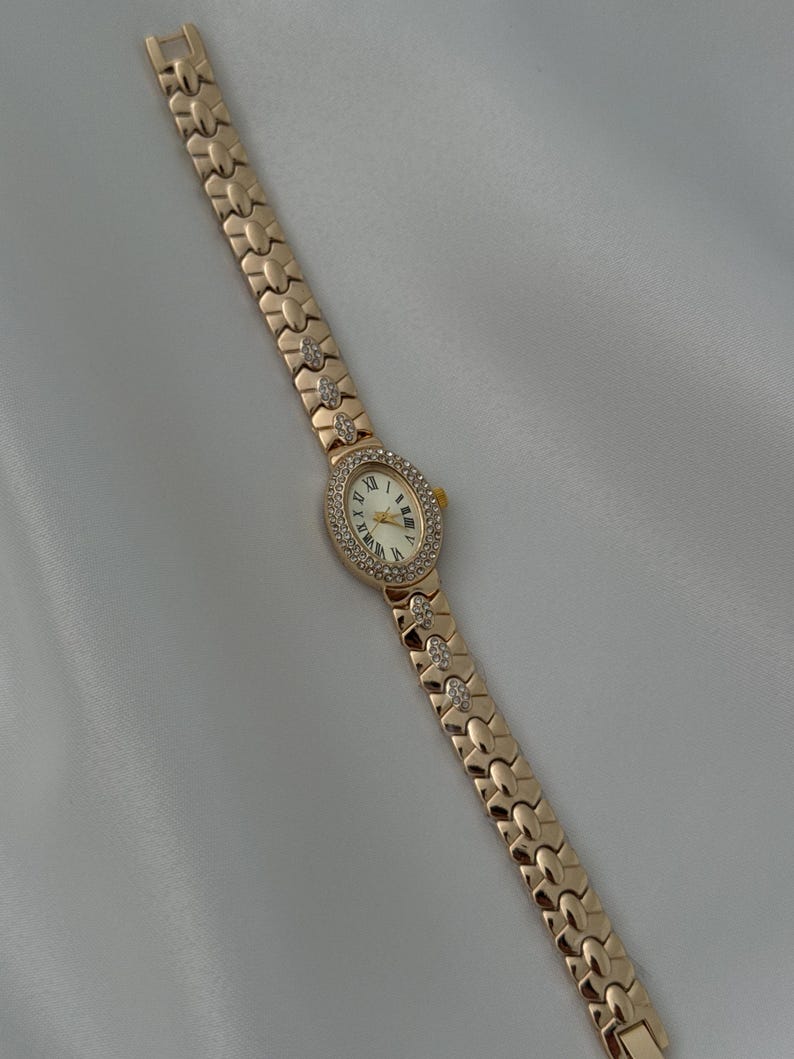 May include: A rose gold-tone wristwatch featuring an oval-shaped face with a white dial, black Roman numeral hour markers, and gold-tone hands. The bezel and select links on the metal bracelet are embellished with small, clear, sparkling stones.