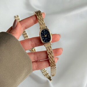 Puede incluir: Reloj de pulsera dorado con esfera ovalada negra y agujas doradas. El reloj tiene una correa de eslabones dorados. El reloj se sostiene sobre un fondo blanco. La esfera del reloj tiene la palabra "QUARTZ" impresa.