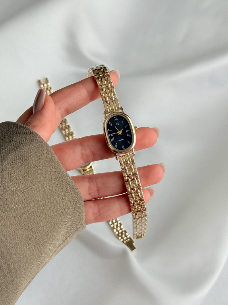 Puede incluir: Reloj de pulsera dorado con esfera ovalada negra y agujas doradas. El reloj tiene una correa de eslabones dorados. El reloj se sostiene sobre un fondo blanco. La esfera del reloj tiene la palabra "QUARTZ" impresa.