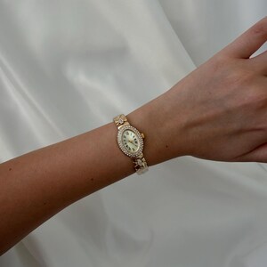 May include: A gold-tone wristwatch featuring an oval-shaped face with a cream-colored dial, Roman numeral markers, and a bezel encrusted with small clear stones. The watch band consists of gold-tone links with decorative stone-set accents.