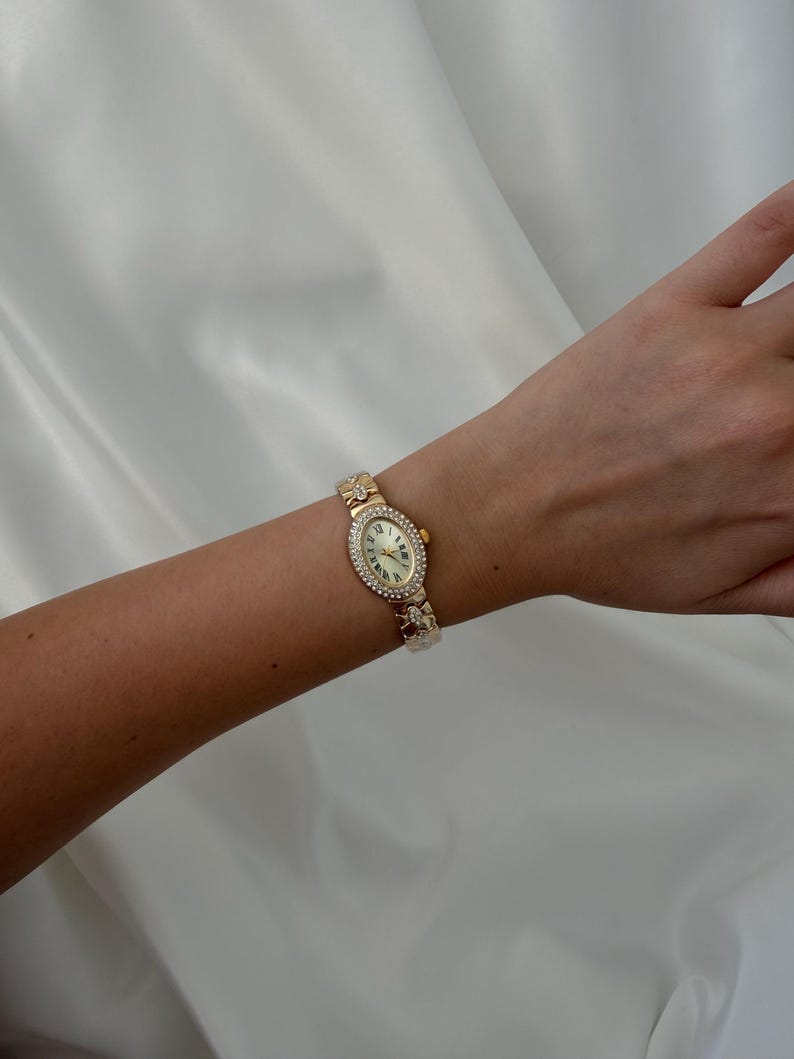May include: A gold-tone wristwatch featuring an oval-shaped face with a cream-colored dial, Roman numeral markers, and a bezel encrusted with small clear stones. The watch band consists of gold-tone links with decorative stone-set accents.