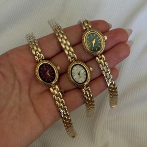 May include: Three gold-tone wristwatches with oval-shaped cases and textured link bands. The watches feature dark red, white, and dark green dials, each with gold-tone hands and a small logo at the twelve o'clock position, all encased in protective plastic.