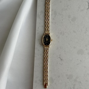 Puede incluir: Un reloj de pulsera dorado con una esfera ovalada negra y n&uacute;meros romanos. El reloj presenta una delicada correa dorada tejida. El reloj se exhibe sobre una superficie de m&aacute;rmol blanco y gris.