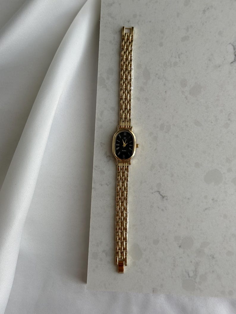 Puede incluir: Un reloj de pulsera dorado con una esfera ovalada negra y n&uacute;meros romanos. El reloj presenta una delicada correa dorada tejida. El reloj se exhibe sobre una superficie de m&aacute;rmol blanco y gris.