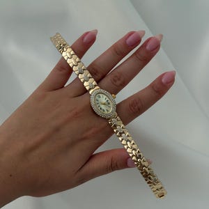 May include: A gold-tone wristwatch featuring an oval-shaped face with a white dial, Roman numeral markers, and a bezel encrusted with small clear stones. The bracelet consists of interlocking gold-tone links, with select links accented by small clear stones.