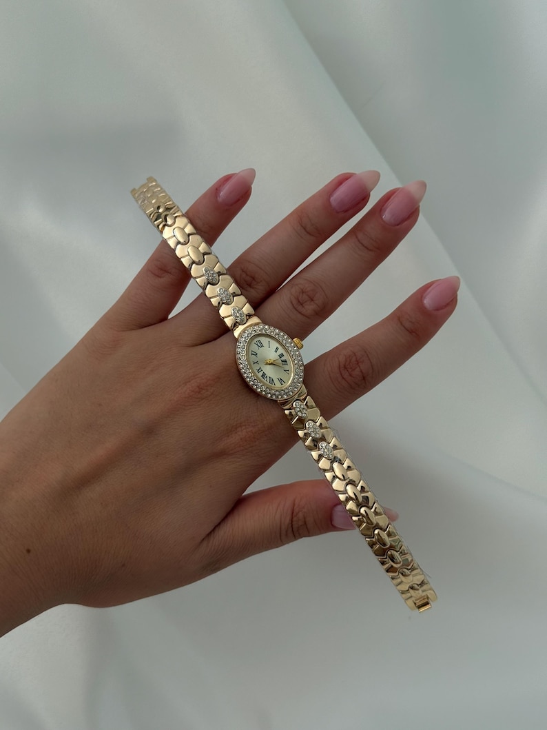 May include: A gold-tone wristwatch featuring an oval-shaped face with a white dial, Roman numeral markers, and a bezel encrusted with small clear stones. The bracelet consists of interlocking gold-tone links, with select links accented by small clear stones.