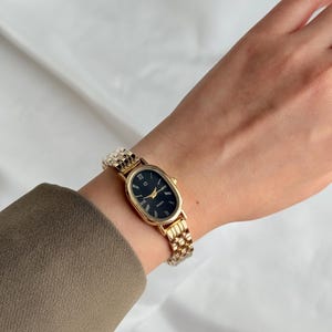 Puede incluir: Reloj de pulsera ovalado dorado con esfera negra y agujas doradas. El reloj presenta n&uacute;meros romanos y una correa de eslabones dorados. El reloj mide aproximadamente 2,5 cm de largo.