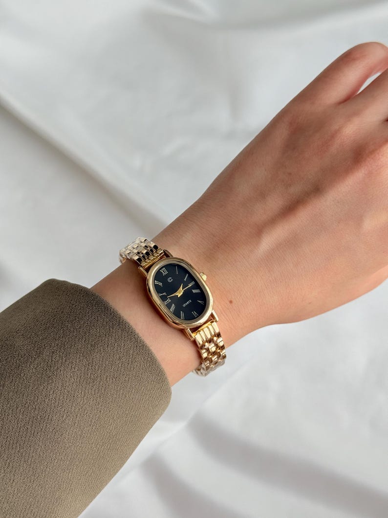 Puede incluir: Reloj de pulsera ovalado dorado con esfera negra y agujas doradas. El reloj presenta n&uacute;meros romanos y una correa de eslabones dorados. El reloj mide aproximadamente 2,5 cm de largo.