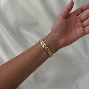 May include: A gold-toned metal link bracelet featuring a textured, nugget-style surface design. The bracelet is constructed with repeating rectangular segments that connect to form a continuous band, secured by a central clasp mechanism.
