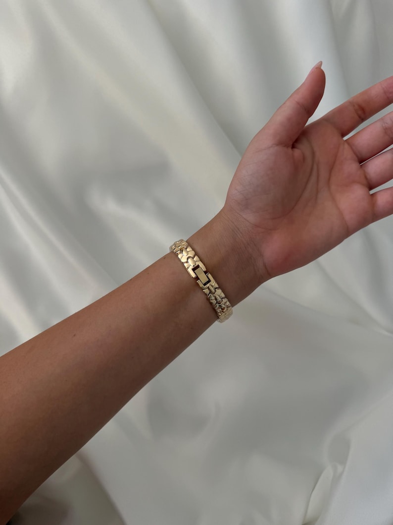 May include: A gold-toned metal link bracelet featuring a textured, nugget-style surface design. The bracelet is constructed with repeating rectangular segments that connect to form a continuous band, secured by a central clasp mechanism.