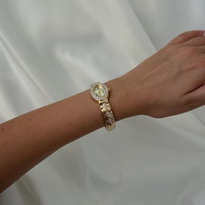 May include: A gold-tone wristwatch featuring an oval-shaped face surrounded by a border of small clear stones. The watch has a metallic link bracelet with decorative cut-out patterns and embedded clear stones along the band.
