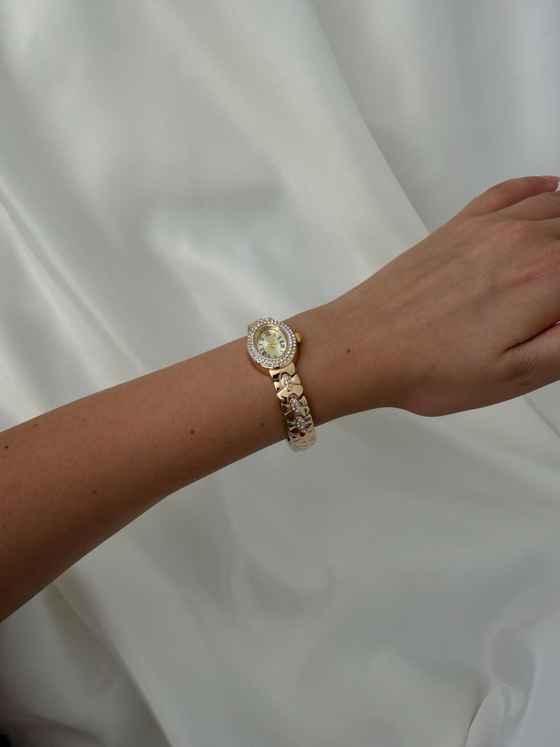 May include: A gold-tone wristwatch featuring an oval-shaped face surrounded by a border of small clear stones. The watch has a metallic link bracelet with decorative cut-out patterns and embedded clear stones along the band.