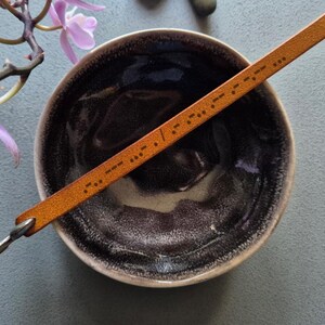 May include: A tan leather bracelet with Morse code lies across a dark brown ceramic bowl. The bracelet has a small metal clasp and a heart-shaped hole at the end. Purple flowers and a grey surface are in the background.