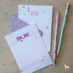 以下が含まれることがあります： 「OH, HI!」と「BW」のピンクと薄紫の文字が入った白いグリーティングカード、ラベンダー色の封筒と紙吹雪。ラベンダー色とミントグリーンの2本の鉛筆がカードの隣にあります。