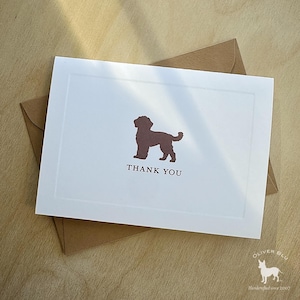 Puede incluir: Una tarjeta blanca con una silueta de perro marrón y las palabras "THANK YOU" impresas en una fuente serif. La tarjeta está dentro de un sobre marrón. La tarjeta está sobre una superficie de madera.