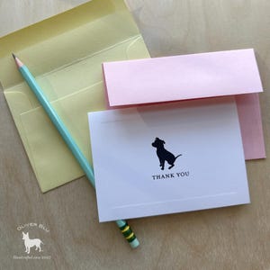 Puede incluir: Un juego de papelería con una tarjeta blanca con una silueta de perro negro y las palabras "THANK YOU". Incluye un sobre rosa claro, un sobre amarillo pálido y un lápiz azul claro. El logotipo de Oliver Blu está en la esquina inferior izquierda.
