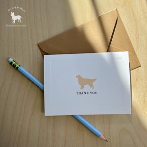 Puede incluir: Una tarjeta de agradecimiento blanca con una silueta de golden retriever y las palabras "THANK YOU" en negro. Un lápiz azul claro y un sobre marrón también están presentes. La tarjeta es parte de un juego de papelería.