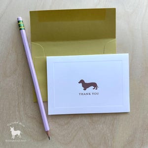 Puede incluir: Una tarjeta blanca con una ilustración de perro salchicha marrón y las palabras "THANK YOU" se coloca frente a un sobre dorado. Un lápiz morado claro con una goma verde descansa en el lado izquierdo de la tarjeta. El fondo es una superficie de madera clara.