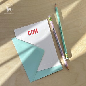 以下が含まれることがあります： 赤い「COH」の文字が入った白いカード、水色の封筒、2本の鉛筆がセットになった文房具セット。鉛筆は、1本は薄い緑色、もう1本はラベンダー色です。鉛筆には黄色と緑のストライプの消しゴムが付いています。