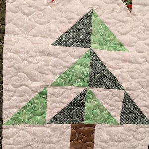 May include: A green and brown patchwork Christmas tree quilt block with a brown trunk. The background is white with a snowflake pattern.