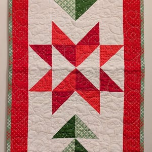 May include: A white quilted table runner with a red border and a green and red Christmas tree design in the center. The tree is made of green triangles and has a brown trunk. The runner is quilted with a snowflake pattern.