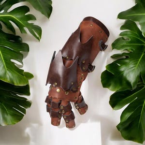 May include: A brown leather gauntlet with a segmented design, featuring dark brown accents and brass-colored rivets. The gauntlet has a spiked edge design and adjustable buckles. The product is displayed against a white background with green leaves.
