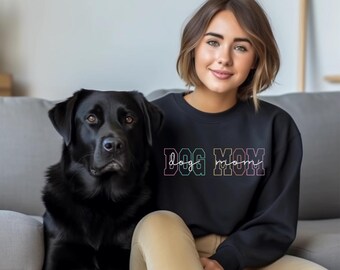 Puede incluir: Una mujer lleva una sudadera negra con el texto "DOG MOM" en colores arcoíris. Está sentada en un sofá con un Labrador Retriever negro.