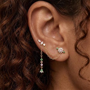 May include: Gold ear cuff with a floral design and dangling chain. The cuff features small, colourful gemstones in green, pink, and white. The chain is adorned with gemstones in various colours, including green and pink. The ear cuff is a stylish accessory.