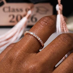 Peut inclure: Bague en or rose ornée de petites pierres précieuses claires. La bague est portée sur un doigt, avec un arrière-plan flou comprenant un gland de remise de diplôme et le texte "Class of 2016."
