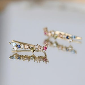 May include: Two gold ear climbers adorned with a variety of colourful gemstones. The earrings showcase a line of gems in shades of blue, pink, white, and green. The earrings are presented on a reflective surface.