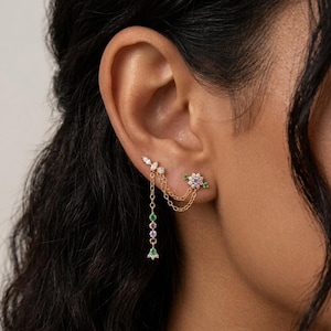Puede incluir: Puño de oreja dorado con un diseño floral y una cadena colgante. El puño presenta un grupo de pequeñas piedras preciosas de colores y una delicada cadena que se conecta a un segundo pendiente con un diseño de flor. Los pendientes se llevan en la oreja de una persona.