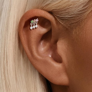 Puede incluir: Un piercing de oreja plateado con un grupo de gemas de colores. La fila superior tiene una gema verde, rosa y transparente. Tres gemas transparentes cuelgan debajo. La joya está colocada en la oreja de una persona.