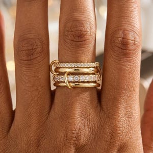 Puede incluir: Primer plano de una mano con varios anillos dorados. Los anillos incluyen una banda lisa, una banda con pequeñas piedras transparentes y otra con piedras más grandes. Un conector dorado une los anillos.