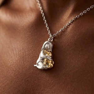 May include: A silver necklace with a pendant depicting a figure embracing a golden-colored animal. The pendant is suspended from a delicate chain. The necklace is a piece of jewelry.