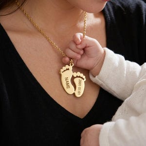 May include: A gold-coloured necklace featuring a pendant shaped like two baby footprints. The pendant is engraved with the word "NAME" and the date "2001 01 01". The necklace is worn around the neck.
