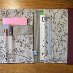 Könnte beinhalten: Ein Notizbuchumschlag aus Stoff mit beigem Hintergrund und einem dunkelbraunen botanischen Zweig- und Beerenmuster. Das Innere enthält Taschen mit einem rosa Notizbuch und zwei Stiften, während rechts ein Schreibblock mit einem weinroten Gummiband befestigt ist.